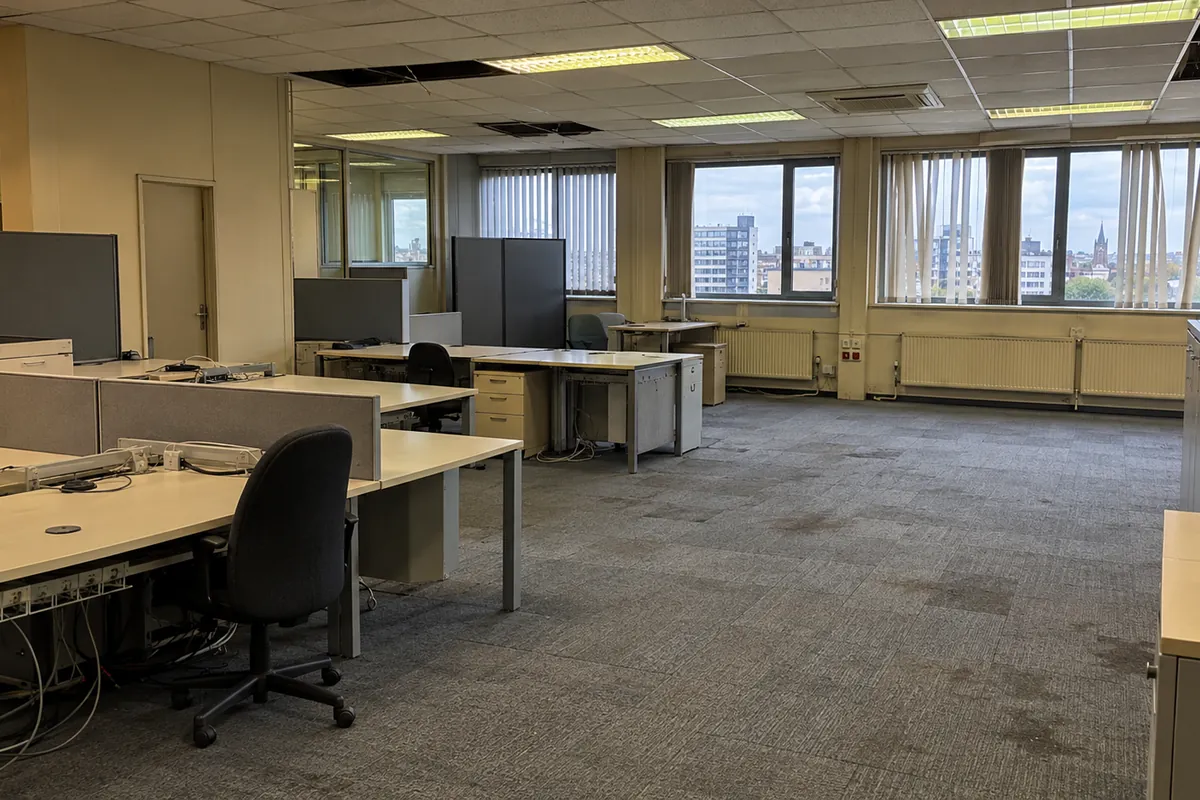 Verouderd open kantoor uit de jaren 90 vóór de renovatie: versleten tapijttegels, systeemplafond met ontbrekende tegels en oude bureaus